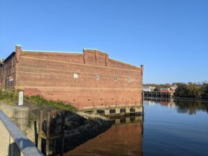 Die Kopfseite eines großen Backsteingebäudes unter blauen Himmel. Es steht auf Holzpfählen im Wasser