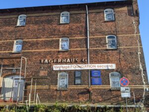 In den Fenstern eines zweistöckigen alten Backstein-Gebäudes hängen fenstergroße schwarz-weiß-Portraits. Unter dem verrosteten Schriftzug "Lagerhaus G" ist ein weißes Transparent mit der Aufschrift "Heritage Foundation secured" aufgehängt.