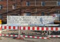 Vor einem Backsteingebäude steht ein weißer Baucontainer. Auf dem ist ein Schriftzug gesprüht (siehe BU), davor eine Baustellenabsperrung