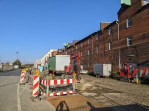 Eine Straße vor einem langgezogenen Backsteinbau ist aufgerissen und mit Baustellenabsperrungen markiert. Bagger und Container stehen herum.