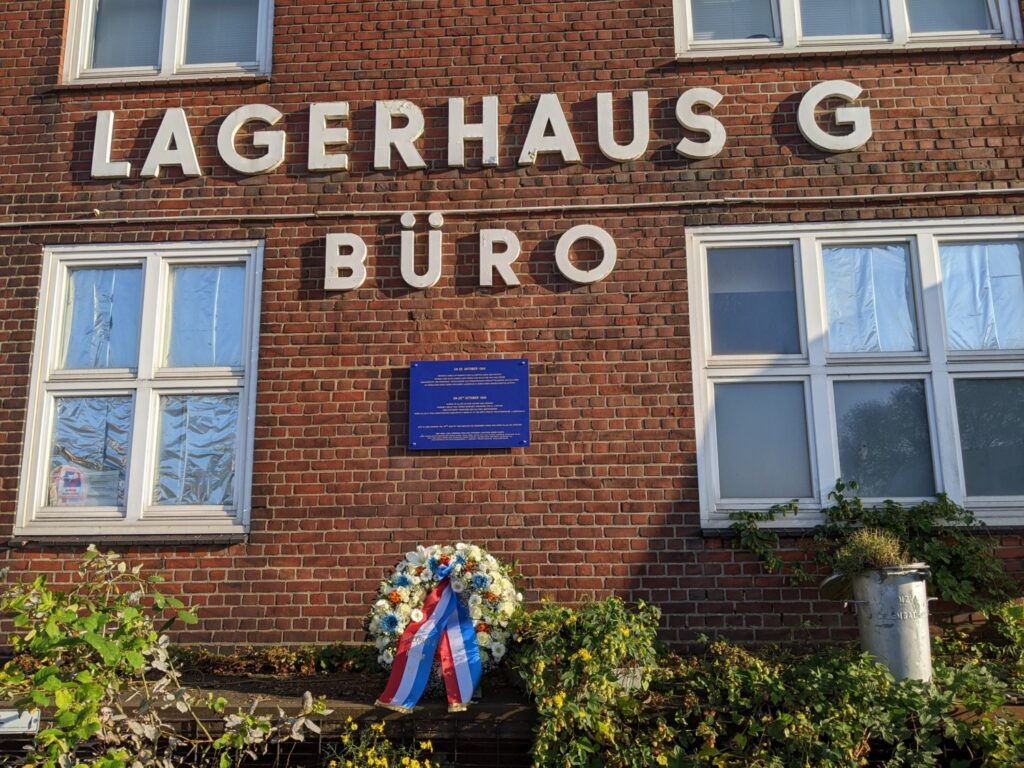 Vor einem Backsteingebäude mit der Aufschrift "Lagerhaus G Büro" unnter der eine kleine, blaue Gedenktafel angebracht ist, steht ein Gedenkkrank auf einem Vorsprung