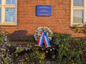 Vor einem Backsteingebäude, an dem eine blaue Gedenktafel hängt, steht ein Trauerkranz, darunter sind Blumen in ein Gitter gesteckt