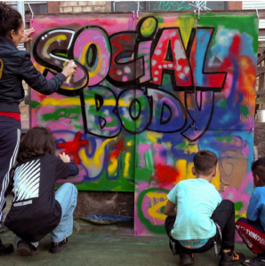 Ein buntes Graffito an einer Wand "Social Body", davor Jugendliche