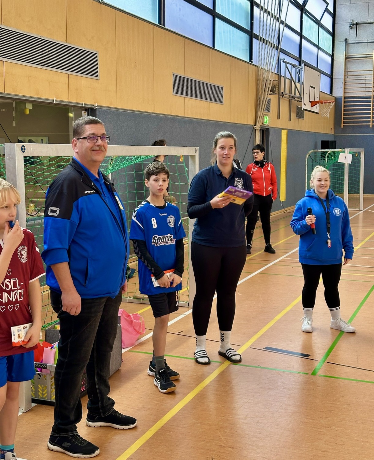 Erwachsene und Kinder in Sportkleidung stehen vor einem Handballtor in einer Sporthalle und halten kleine Geschenke in den Händen.