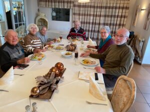 zwei Damen und vier Herren sitzen um einen Tisch mit einer broncefarbenen Deko und eicherschalenfarbenen Tischdecke und Servietten. Sie haben Teller mit Essen und Getränke vor sich und lächeln in die Kamera.