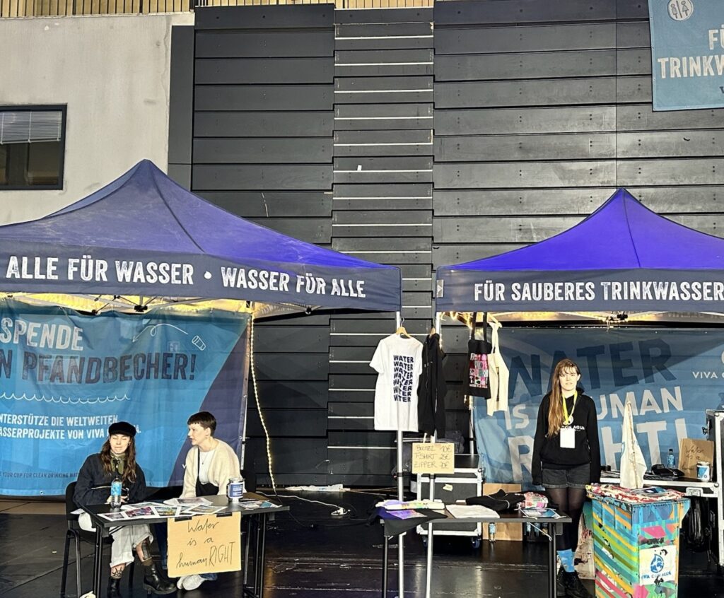 Zwei blaue Pavillon-Dächer mit den Aufschriften "Alle für Wasser, Wasser für Alle" und "Für sauberes Trinkwasser", darunter Menschen an Ständen mit Artikeln