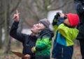 Ein Erwachsener und Kinder in Winterkleidung, Bäume im Hintergrund, sehen nach oben, ein Kind hält ein Fernglas vor die Augen