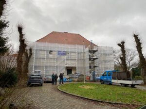 In der Mitte ein großes Gebäuse mit einer Plane vor der Fassade. Rechts und links kahle Bäume. Vor dem haus links 2 PKWs, daneben 3 Leute, dann ein Müllcontainer und rechs ein Lastwagenl