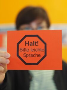 Eine Person im Hintergrund hält ein Schild in der Hand. Es ist orange, darauf der Umrss eines Stoppschilds. Darauf steht: Halt! Bitte leichte Sprache