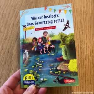 Ein Pixie-Buch auf einem Tisch, das ene Hand festhält. Titel des Buchs: Wie der Inselpark Opas Geburtstag rettete