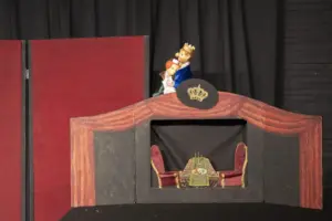 Das Bild zeigt eine Puppentheater-Bühne mit einer königlichen Szenerie. Im oberen Bereich sind zwei Puppen zu sehen: ein König mit goldener Krone und blauer Robe, der eine Königin oder Prinzessin in den Armen hält. Die Bühne ist mit einem gemalten roten Vorhang und einem goldenen Kronensymbol verziert. Im unteren Bereich der Bühne ist eine kleine königliche Tafel mit zwei roten Sesseln, einem gedeckten Tisch mit Speisen und einem Kerzenständer dargestellt. Der Hintergrund besteht aus schwarzen Vorhängen und roten Stoffpaneelen, was eine Theateratmosphäre erzeugt.