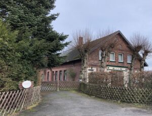 Hinter einem Zaun mit Toreinfahrt steht ein altes Backsteingebäude, das an ein Bauernhaus erinnert. Es wirkt verlassen und verriegelt. Eine hohe Hecke, Tannen und Weiden sind auf dem Grundstück zu sehen.