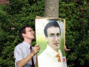 Ein Mann, schlank, kurze Haare, Brille, Hemd und Schlipps, hänge ein Wahlplakat von ihm selbst an einem Baum 