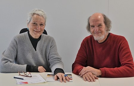 Links im Bild eine Seniorin im grauen Pullover, rechts im Bild ein Senior im roten Pullover, die an einem Tisch sitzen und freundlich lächelnd in die Kamera schauen