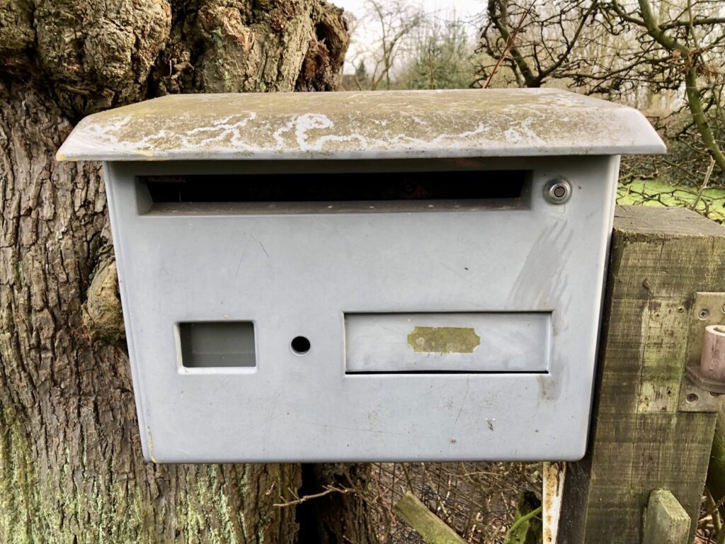 ein verlassen wirkender Briefkasten am Zaunpfahl, im Hintergrund ein Baumstamm