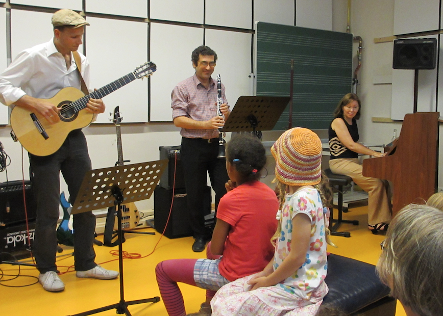 Im Musikatelier spielen von links ein Gitarrist, ein Klarinettist und eine Pianistin. Im Vordergrund die Rückansicht von zwei kleinen Mädchen, die zuhören