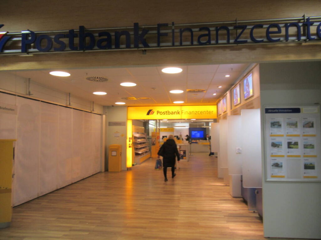 Am Ende eines Ganges befindet sich noch die Postbank im Luna-Center. Sie wird mit einem gelben Leuchtschild am Eingang beworben. Man sieht auch einen Briefkasten, einen Bildschirm, ein Regal mit Briefumschlägen und einen Menschen, der auf die Filiale zugeht.