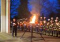 Links eine hohe weiße Saäule. Rechts daneben Feuerwehrleute mit Fackeln. Vor der Säule ein Mann und in der Mitte eine große Feuerschale