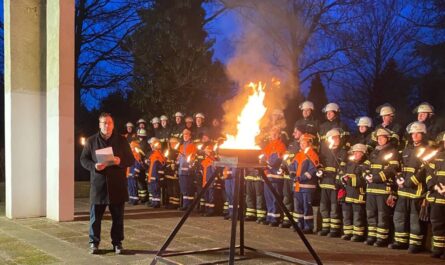 Links eine hohe weiße Saäule. Rechts daneben Feuerwehrleute mit Fackeln. Vor der Säule ein Mann und in der Mitte eine große Feuerschale