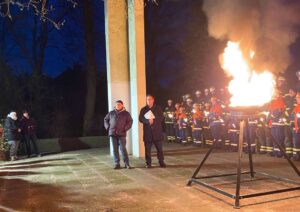 Vor einer hohen weißen Säule in der Mitte stehen zwei Männer. Im Hintergrund rechts viele Feuerwehrleute mit Fackeln. Rechts eine Feuerschale auf drei Beinen mit einem großen Feuer.