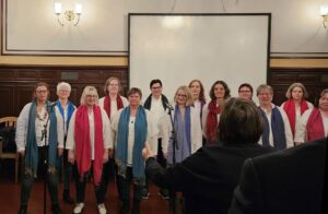 Viele Frauen mit unterschiedlich farbigen langen Schals stehen vor einem Flipchart und einer Wand mit Wandleuchtern.