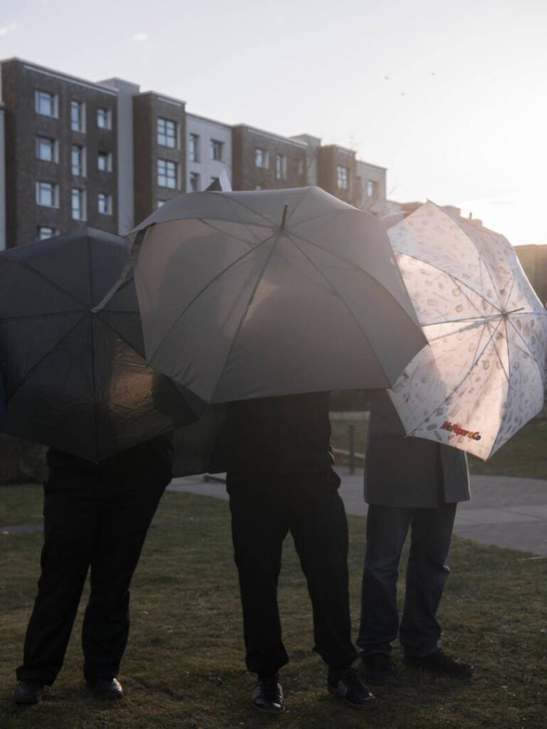 Im Vordergrund Menschen, die Regenschirme vor dem Gesicht aufgespannt haben, im Hintergrund Mehrfamilienhäuser