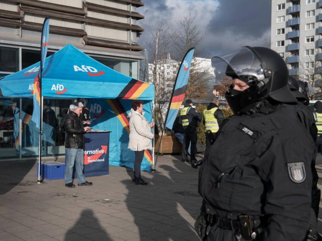 Vor einem AfD-Pavllon stehen eine Frau mit kurzen roten Haaren und ein Mann mit Fellmütze, davor ein behelmter Polizist