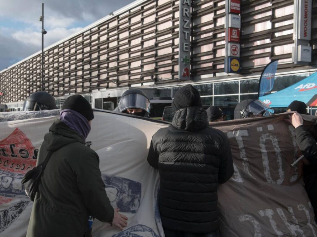 Menschen von hinten, die ein Transparent tragen, ein behelmter Polizist, der wütend durch sein Versier guckt