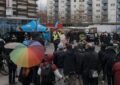 Vor Hochhäusern ist im Grund ein blauer Pavillon mit der Aufschrift AfD zu erkennen, davor stehen Polizist*innen, im Kreis darum viele Menschen, einige davon mit Regenschirmen