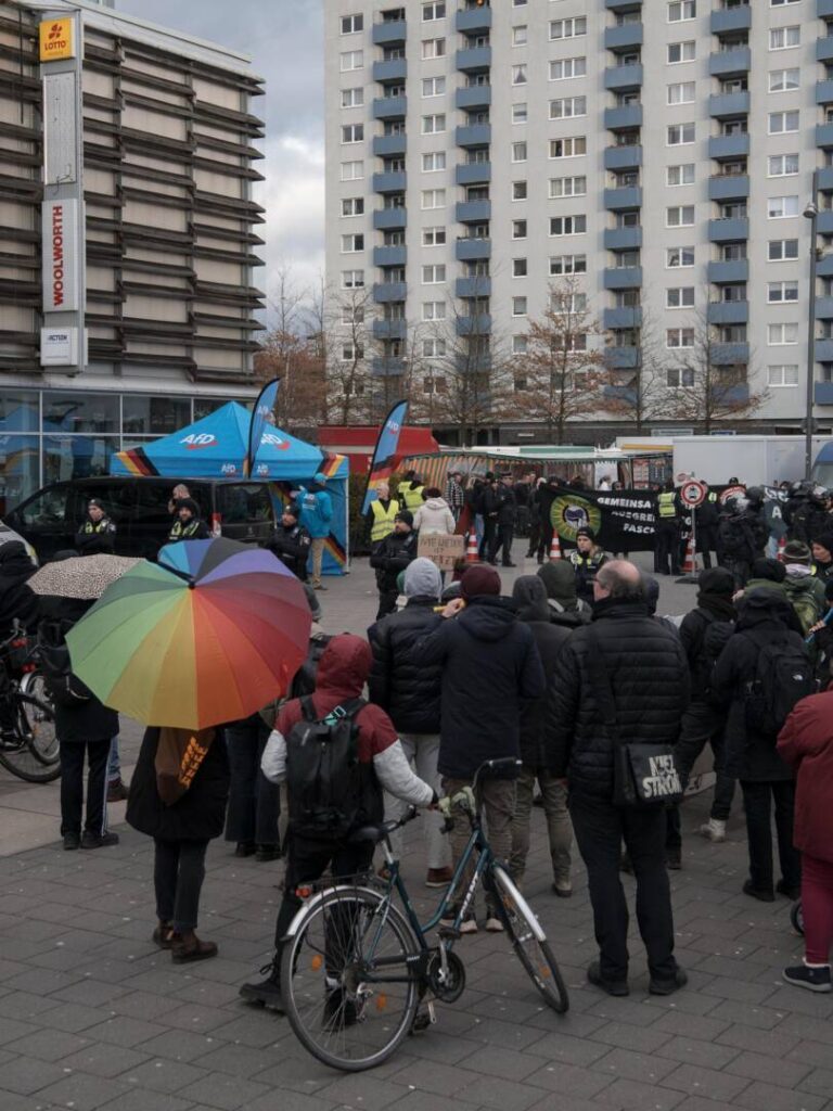 Vor Hochhäusern ist im Grund ein blauer Pavillon mit der Aufschrift AfD zu erkennen, davor stehen Polizist*innen, im Kreis darum viele Menschen, einige davon mit Regenschirmen