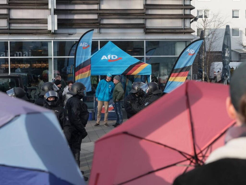 Vor einem AfD-Pavillon stehen Polizist*innen in Kampfmontur, davor sind aufgescpannte Regenschirme zu sehen