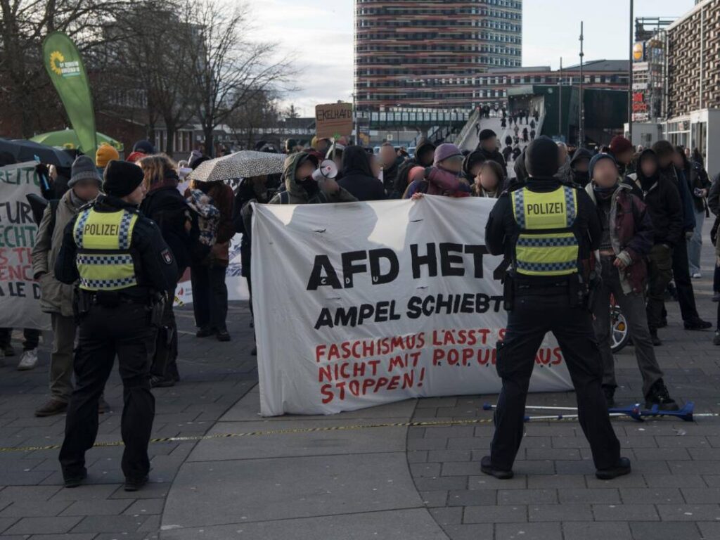 Hinter einen großen Transparent auf dem steht: "AfD hetzt, Ampel schiebt ab" stehen Demontrierende, davor ein Polizist in gelber Warnweste