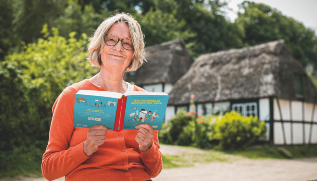 Die Autorin Kirsten Boie sitzt mit aufgeschlagenem Buch mit blauem Layout. Sie lächelt in die Camera, trägt eine Brille und einen oangenen Pulli. Im Hintergrund sind Bäume, Büsche, ein Weg und 2 Fachwerkgebäude zu sehen.