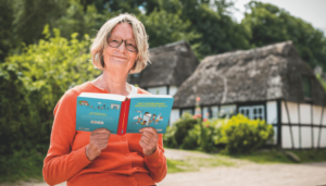 Die Autorin Kirsten Boie mit aufgeschlagenem Buch mit blauem Layout. Sie lächelt in die Kamera, trägt eine Brille und einen oangenen Pulli. Im Hintergrund sind Bäume, Büsche, ein Weg und 2 Fachwerkgebäude zu sehen.