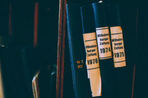 Das Bild zeigt eine Nahaufnahme eines Bücherregals mit mehreren gebundenen Ausgaben der „Wilhelmsburger Zeitung“ aus den Jahren 1974, 1975, 1976 und 1977. Die Buchrücken sind schwarz, mit weißen oder goldenen Beschriftungen. Die Jahrgänge 1974 bis 1976 haben abgenutzte weiße Papieretiketten mit schwarzem Text, während die Ausgabe von 1977 mit goldenen Buchstaben „W Z 1977“ direkt auf den Buchrücken geprägt ist. Die Bücher stehen dicht nebeneinander in einem dunklen Regal, das eine nostalgische, historische Atmosphäre vermittelt.