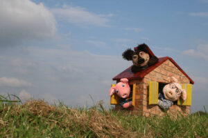 Das Bild zeigt eine Szene mit drei Handpuppen in einem kleinen Spielhaus auf einer Wiese unter blauem Himmel mit einigen Wolken. Zwei Schweinepuppen schauen fröhlich aus den gelben Fensterläden des Hauses – eines davon trägt eine Brille und ein gestreiftes Shirt, das andere ein dunkles Hemd mit Muster. Auf dem Dach des Hauses liegt eine Hundepuppe mit schwarzen Ohren und einem breiten Grinsen. Das Haus besteht aus gemustertem Karton, der wie Ziegel aussieht, und hat ein rotes Dach.