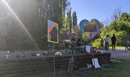 Auf einer hölzernen Parkbank liegen Blumen und stehen Kerzen, davor 2 Schilder an Stecken. Auf dem einen reckt sich eine geballte schwarze Faust in die Luft, auf dem anderen steht: "If you are not working for justice stop calling for peace"