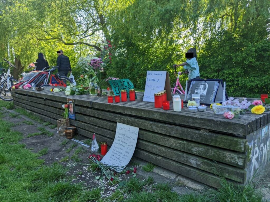 Im Hintergrund grüne Weiden , vorbeigehende Menschen, ein Kind auf einem Fahrrad. Mittig eine lange Holzbank, auf der Windlichter, Fotos und Blumen stehen und liegen. Davor ein Schild mit den Namen getöteter Schwarzer Menschen der letzten Jahre in Deutschland und weitere Blumen und Kerzen