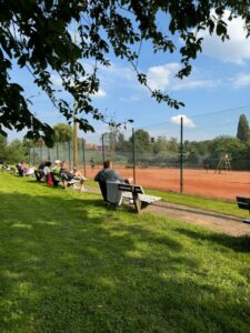 unterschiedliche Menschen sitzen auf mehreren Bänken bei schönem Wetter am Tennisplatz