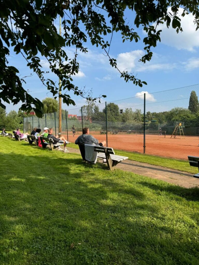 unterschiedliche Menschen sitzen auf mehreren Bänken bei schönem Wetter am Tennisplatz