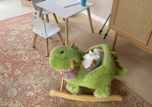 Kinderspielecke mit kleinem Tisch und zwei Kinderstühlen. Rechts die Ecke von einem Sideboard und davor auf einem Teppich ein grünes Plüschtier, auf dem noch ein Plüschigel sitzt.