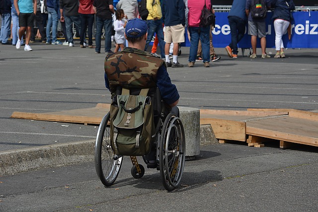 Rückansicht eines Mannes im Rollstuhl. Um ihn herum sind Poller und eine Bordsteinkante aus Beton, davor befindet sich noch eine Holzrampe, die quer zu seiner Fahrtrichtung aufgebaut ist. Er ist also von HIndernissen umgeben und kann deshalb nicht zu den anderen Menschen, die alle gehen können, hinkommen.