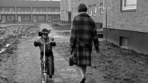 Schwarz-weiß-Foto: Ein Kind auf einem Roller von vorne und eine Frau von hinten im Mantel auf einem Weg vor Häusern