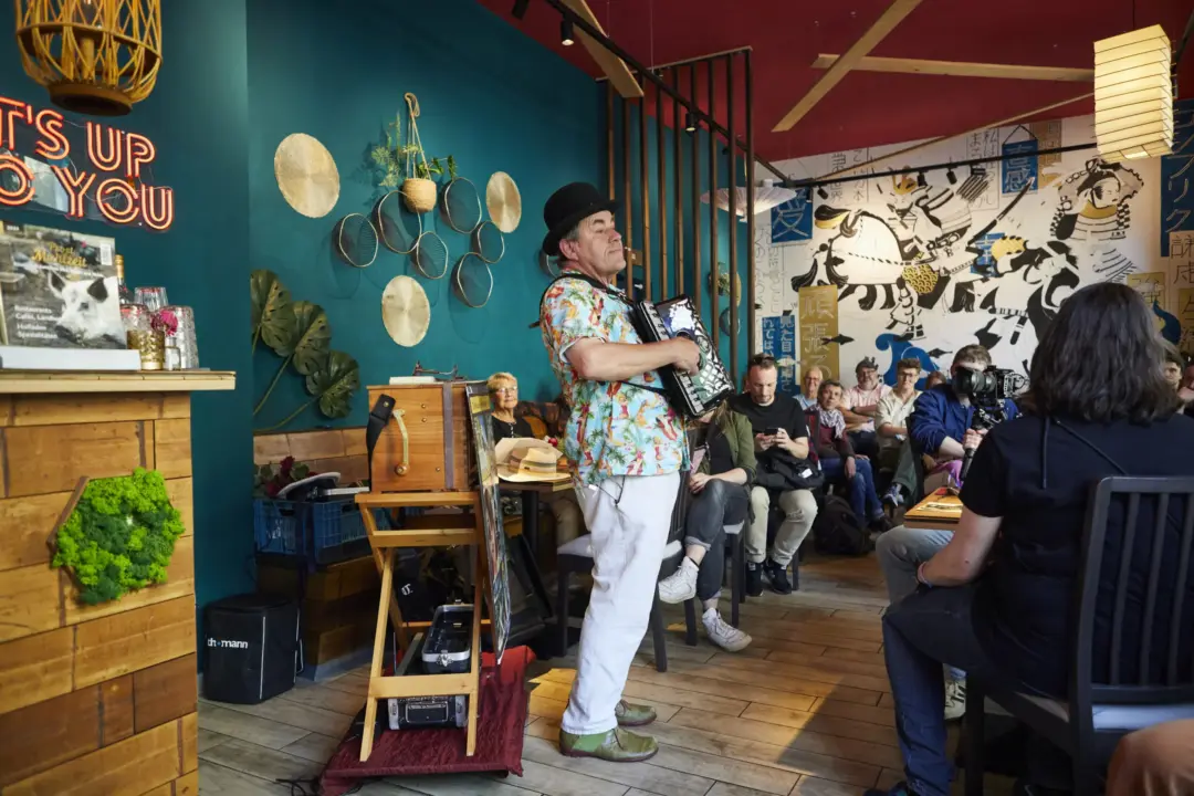 Ein Musiker mit Akkordeon und Melone spielt im bunt gestalteten Restaurant "Aomame". Er trägt ein farbenfrohes Hawaiihemd, weiße Hose und grüne Schuhe. Links steht eine Drehorgel mit einem offenen Kasten und technischem Zubehör davor. Im Hintergrund sitzt ein aufmerksames Publikum auf Bänken und Stühlen, einige filmen oder fotografieren. Die Wände sind kreativ gestaltet: eine Seite mit dekorativen Körben und Pflanzen, die andere mit großflächiger Wandmalerei asiatischer Motive. Über dem Tresen leuchtet ein Neon-Schriftzug: „IT’S UP TO YOU“.
