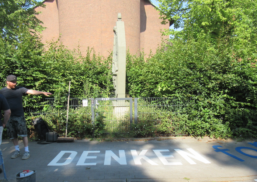 Das gedrehte Denkal von der Straße aus betrachtet. Davor auf dem Gehweg in thermoplastischen Großbuchstaben das Wort denken