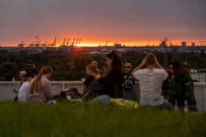 Menschen sitzen auf einer Wiese auf einem Berg und schauen auf eine Aussicht mit Hafen und Sonnenuntergang