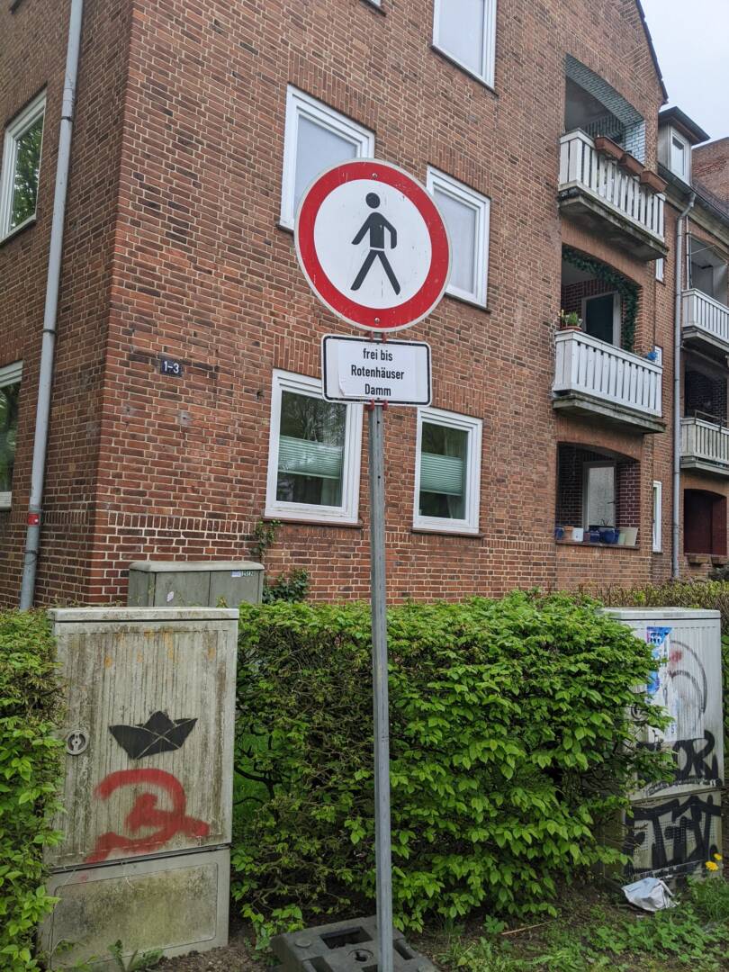 Ein rundes Schild mit rotem Rand, darin ein Fußgänger-Männchen. Im Hintergrund ein Backsteingebäude