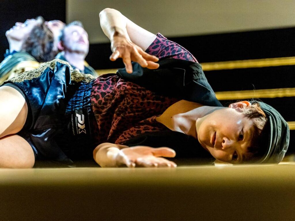 Ausschnitt aus dem Theaterstück: eine Frau liegt mit angewinkelten Beinen auf der Seite und gestikuliert mit ihren angewinkelten Armen und Händen. Sie trägt kurze dunkle Haare, ein Cappy sowie kurze shorts und ein shirt im leoparden ähnlichem muster in rot-schwarz mit einer schwarzen Weste drüber. Im Hintergrund sind aneinandergelehnte Köpfe von drei weiteren Personen zu erkennen, die im scheinwerferlicht sitzen