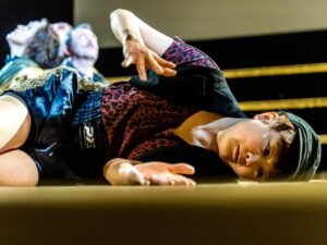 Ausschnitt aus dem Theaterstück: eine Frau liegt mit angewinkelten Beinen auf der Seite und gestikuliert mit ihren angewinkelten Armen und Händen. Sie trägt kurze dunkle Haare, ein Cappy sowie kurze shorts und ein shirt im leoparden ähnlichem muster in rot-schwarz. Im Hintergrund sind aneinandergelehnte Köpfe von drei weiteren Personen zu erkennen, die im scheinwerferlicht sitzen