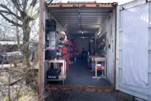 Neben einem Baum steht rechts ein Container mit offener Tür, in dem rechts und links an den Seiten Werkzeug gelagert wird.